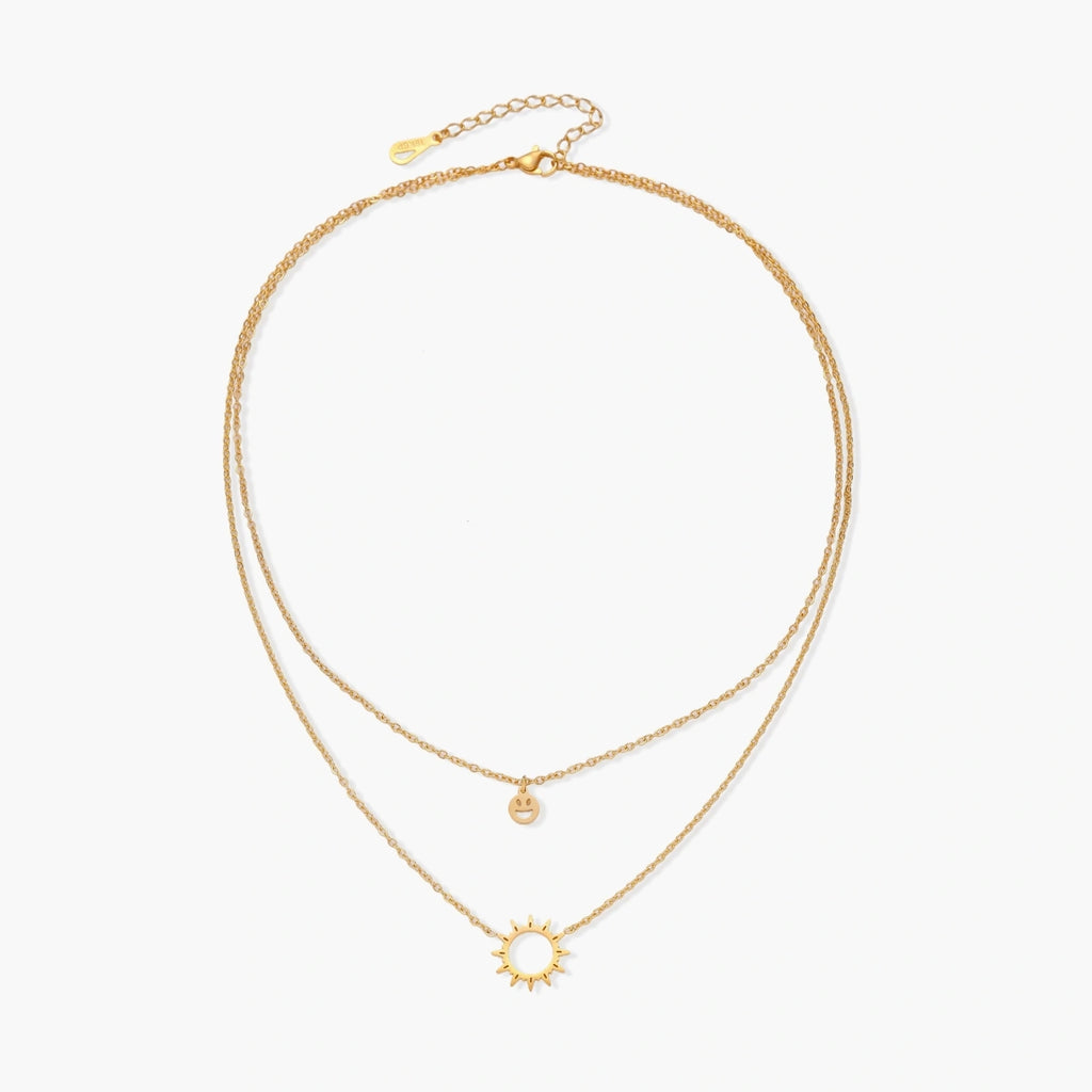 Gold layered necklace set with sunburst and smiley face pendants on delicate chains against white background.
